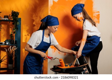 Woman And Girl Renovating House With Paint, Using Orange Color To Decorate Wall Surface In Apartment. Young Child On Ladder Painting With Paintbrush And Adult With Rolling Brush, Having Fun.