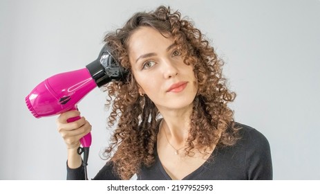 Woman gives herself a curly hairstyle. Hair care concept. A curly woman dries her hair at home with a hair dryer with a diffuser attachment. A beautiful girl uses a modern hair dryer