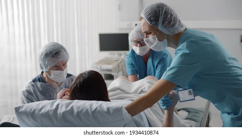 Woman giving birth with husband holds her hand in support and obstetricians assisting. Back view of medical staff in protective uniform helping pregnant woman in labor