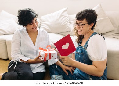 Woman Giving A Gift And Reading A Card For Her Girlfriend At Home. Lesbian Couple With Gift And Valentines Card.