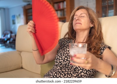 Woman Giving Herself Air With A Fan And A Glass Of Water With Ice To Cool Off In Summer.