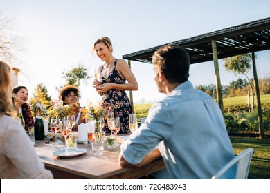 Woman Giving A Speech To Friends At Outdoors Party. Young People Celebrating A Special Occasion In A Garden Restaurant.