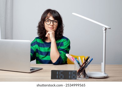 A woman in glasses and a green-striped shirt sits thoughtfully at a desk, her chin resting on her hand. A laptop, pens, and a digital clock showing 16:16 are arranged neatly on the desk. - Powered by Shutterstock