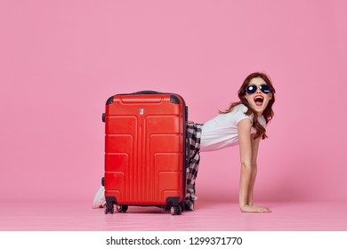 A Woman In Glasses Is Kneeling Behind A Red Suitcase On A Pink Background                     