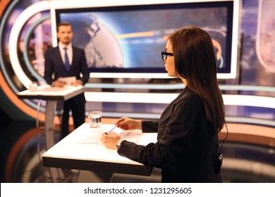 Woman In Glasses And Man In Suit Standing In Studio, Making A TV News Show. A Glass Of Water And Paper Leaf On The Table. Big Screen On The Background.