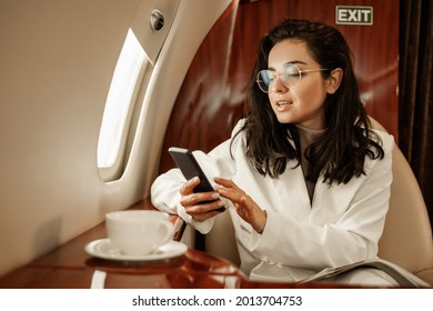 A Woman With Glasses On The Phone Checks Her Social Networks During A Coffee Break In The Flight Of A Private Plane. Travel After Covid Pandemic