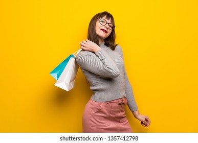 Woman with glasses over yellow wall holding a lot of shopping bags