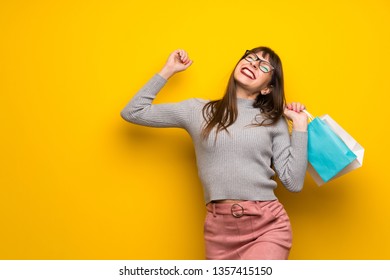 Woman with glasses over yellow wall holding a lot of shopping bags in victory position