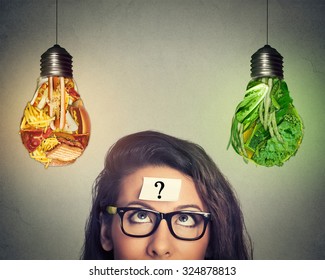 Woman in glasses question mark on head thinking looking up at junk food and green vegetables shaped as light bulb isolated on gray background. Diet choice right nutrition healthy lifestyle concept   - Shutterstock ID 324878813