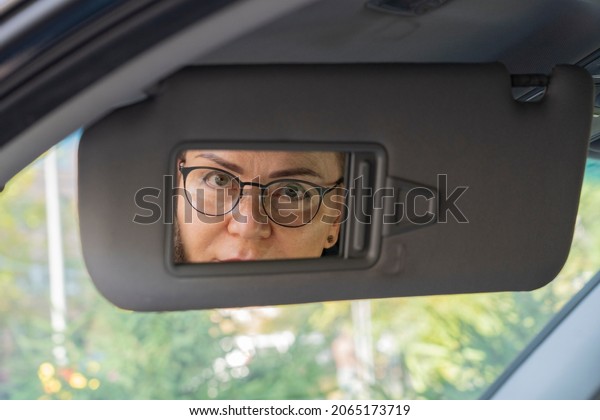 A woman with glasses sits in a car and looks in\
the mirror.
