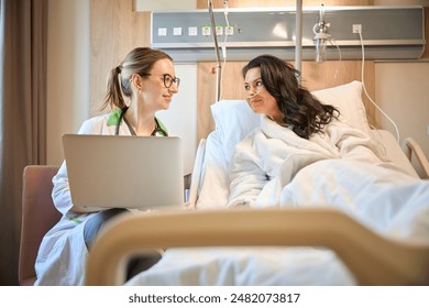 Woman with glasses sits with laptop near lady on hospital bed - Powered by Shutterstock