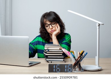 A woman in glasses and a striped green and navy sweater sits at a desk, looking bored and resting her head on her hand. She is surrounded by a laptop, a stack of books, pens, and a digital clock.  - Powered by Shutterstock