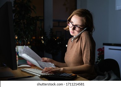 Woman In Glasses Using Calculator To Calculate The Invoice, Planning Expenses, Talking On Smartphone, Working On Desktop Pc Late At Night At Home Office, Can Not Sleep. Family Budget, Remote Work. 