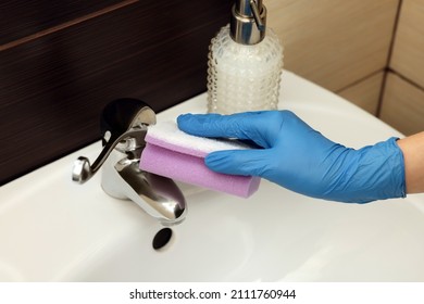 Woman in gloves cleaning faucet of bathroom sink with sponge, closeup