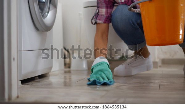 Woman in gloves having leaking\
washing machine wiping floor and wrinkling rag in bucket. Housewife\
near broken washing machine collecting water in basin in\
bathroom