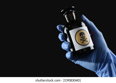 Woman In Gloves Holding Glass Bottle Of Poison With Warning Sign On Black Background, Closeup. Space For Text