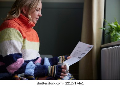 Woman In Gloves With Hot Drink And Bill Trying To Keep Warm By Radiator During Cost Of Living Energy Crisis - Shutterstock ID 2212717461