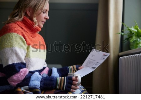 Woman In Gloves With Hot Drink And Bill Trying To Keep Warm By Radiator During Cost Of Living Energy Crisis Foto stock © 