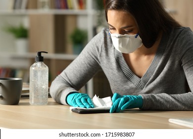 Woman with gloves and protective mask disinfecting smart phone from coronavirus sitting on a desk at night at home