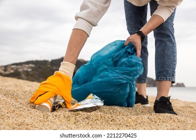 A Woman In Gloves With A Special Blue Bag Picks Up Garbage Among The Sand Along The Coast. The Problem Of Environmental Pollution. Cleaning Up Trash On The Beach