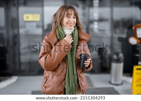 Woman going out from shop or small supermarket with coffee cup at gas station. Young woman wearing face mask and dressed in warm clothes. Buying take away drink during pandemic
