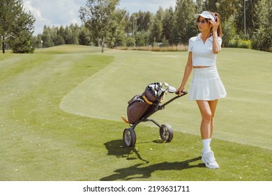 Woman golf player walking on fairway with golf trolley. Wearing white sporty uniform. Short skirt, polo and cap - Powered by Shutterstock