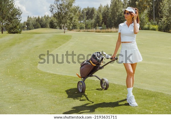 Woman golf\
player walking on fairway with golf trolley. Wearing white sporty\
uniform. Short skirt, polo and\
cap