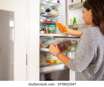 Woman Grabbing Carrots From The Drawer Fridge For Cooking A Healthy Salad. Young Female Searching Food In Open Refrigerator With Leftovers Container. Meal Preparation For Dieting.