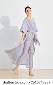 Una mujer posa con gracia en un vestido de lavanda que fluye con una división, destacando su estilo elegante. Se encuentra en una habitación bien iluminada con paredes blancas