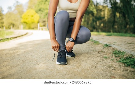 A woman in gray leggings and a white tank top sits on a dirt path in a park, tying her running shoes. She is wearing a smartwatch on her wrist, cropped - Powered by Shutterstock