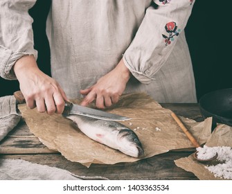 Woman In Gray Linen Clothes Cleans The Fish Sea Bass Scales On A Brown Wooden Board, Black Background