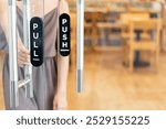 A woman in a gray outfit is interacting with glass doors that have pull and push signs. The setting appears to be a cafe or restaurant with wooden furniture in the backer