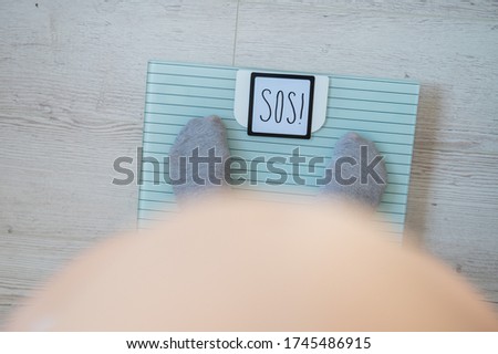 A woman in gray socks is standing on an electronic scale. Top view on the legs and bare belly of an obese woman. The word sos on the scales display.