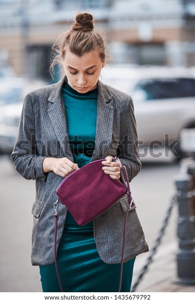 woman in a green dress and a gray\
jacket opens her bag, against the background of the\
city
