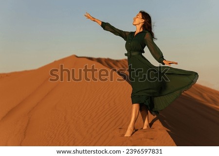 woman in green long dress stands in desert sand dunes background yellow orange sunset