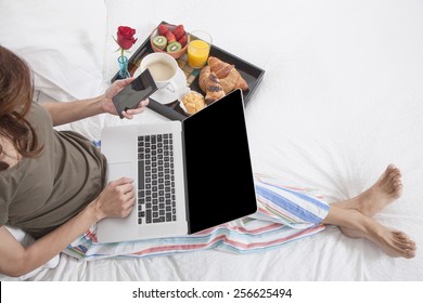 woman green shirt striped pajama pants sitting on white bed with laptop and mobile phone breakfast tray croissants orange juice strawberry kiwi cupcake red rose flower