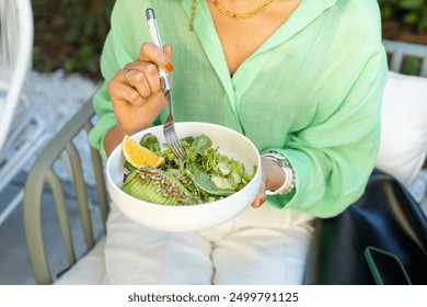  Woman in green t-shirt and jeans holding vegan superbowl with hummus, vegetable, salad, beans, couscous and avocado. Healthy dinner or lunch