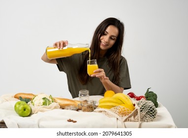 Woman in green t-shirt pouring a juice from a glass bottle in a glass over a table with mesh eco bag, healthy vegan vegetables, fruits, bread, snacks. Zero waste concept. - Powered by Shutterstock