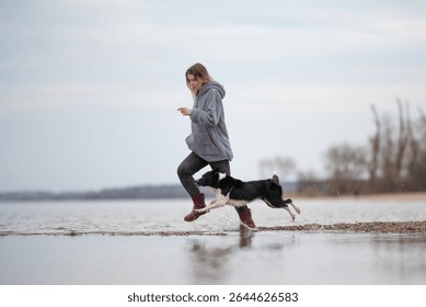 A woman in grey clothing and red boots runs alongside a black and white border collie dog. They splash through the shallow water near the shoreline on an overcast day