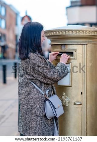 woman in grey coat posting letters in gold post box in England.  Olympic post box