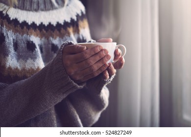 Woman in grey knitted sweater with traditional motifs holding in hands a white cup of hot coffee with steam coming out in the morning light