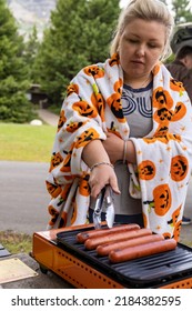 Woman Grills Hot Dogs On An Outdoor Camp Stove Using Tongs, At A Picnic Site