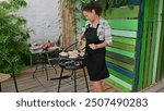 A woman grills seafood on a barbeque in a garden, enjoying outdoor cooking amidst greenery and a colorful patio setting.