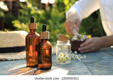 Woman Grinding Chamomile Flowers In Mortar Outdoors, Focus On Bottles Of Essential Oil