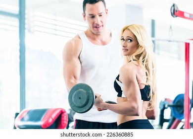 Woman In Gym Training For Body Building Competition With Barbell Assisted By Personal Fitness Trainer