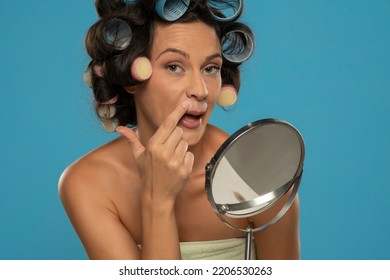 Woman With Hair Curlers Applies Concealer On Her Upper Lips On A Blue Studio Background