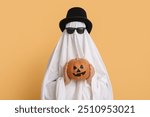 Woman in Halloween costume of ghost, cylinder hat and sunglasses holding pumpkin on color background