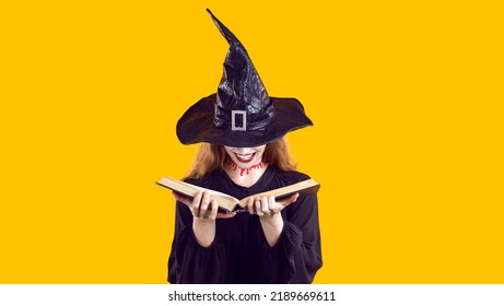 Woman In Halloween Costume Looking Through Book Of Spells. Cunning Evil Witch With Bloody Neck Cut And Creepy Happy Smile Hides Face Under Hat And Chooses Most Terrible Curse From Witchcraft Manual