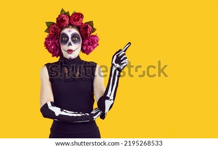 Woman in Halloween costume with surprised expression showing copy space advert solution. Woman in peony wreath, Halloween makeup and gloves in form of hand brush advertises on orange background.