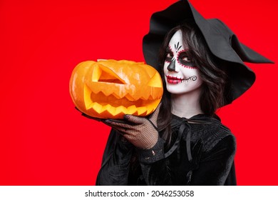 Woman in Halloween costume wear lace gloves, black witch hat, with creative sugar skull makeup. On red background hold magic scary pumpkin. Ð¡oncept Los Muertos poster party or La Calavera Catrina.